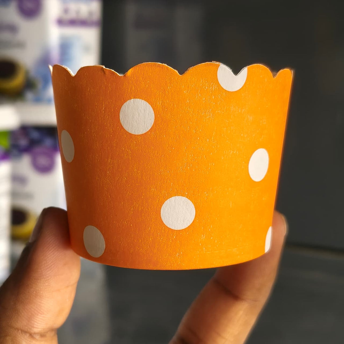 An orange paper cupcake liner with white polka dots, held by a hand against a blurred background.