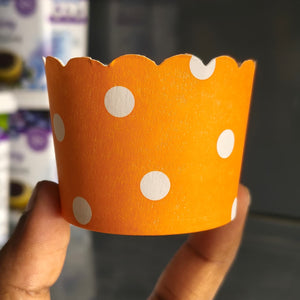 An orange paper cupcake liner with white polka dots, held by a hand against a blurred background.