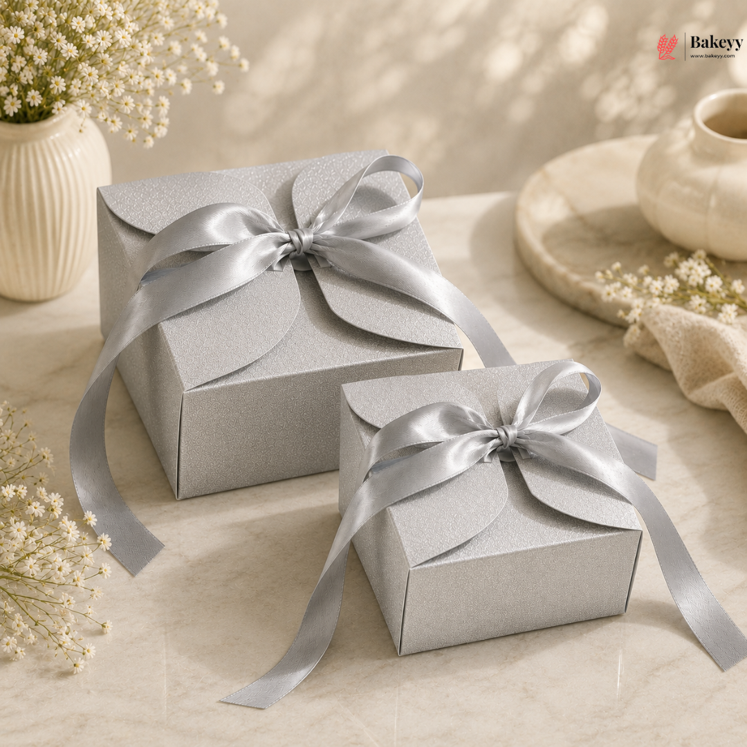 Two silver gift boxes with ribbons on a light surface with flowers and a teapot in the background.
