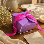 Purple gift box with pink ribbon on a wooden surface, surrounded by flowers and a card.