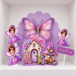 A purple fairy-themed birthday party decoration set featuring a large butterfly, two fairies, a flower-covered fairy house, a small mushroom house, and a 'Happy Birthday' sign, displayed on a white shelf.