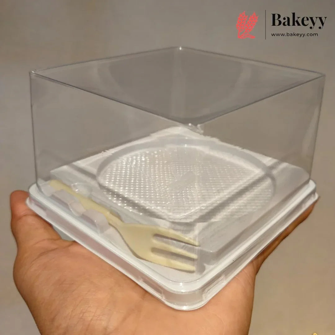 A hand holding a clear plastic bakery display box with a white base and a small beige plastic fork inside, against a plain light background. The Bakeyy logo is visible in the top right corner.