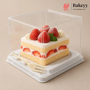 A clear square plastic cake display box with a white base, shown containing a slice of strawberry cake with cream and fresh strawberries on top. A small cream fork rests inside the box.