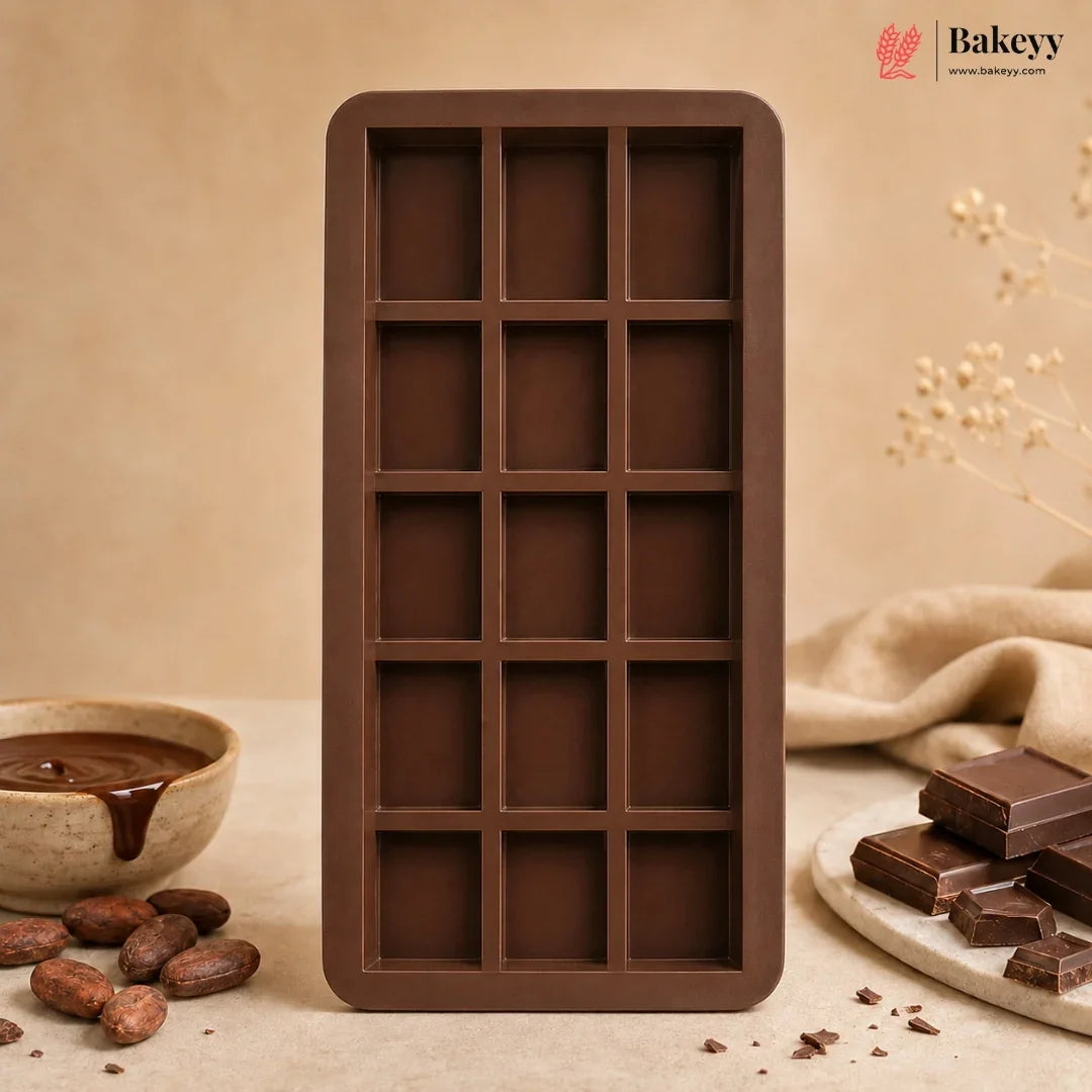 A rectangular chocolate mold with 15 dark brown cavities, styled on a beige surface with chocolate pieces, cocoa beans, and a bowl of chocolate sauce. The Bakeyy brand logo is visible in the top right corner.
