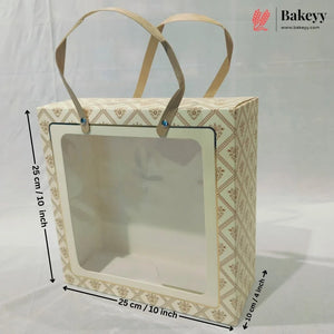 A square bakery gift box with a clear front window and beige paper handles, shown with dimension arrows indicating 20 cm by 20 cm by 10 cm measurements on a white backdrop. A Bakeyy logo appears in the top right.