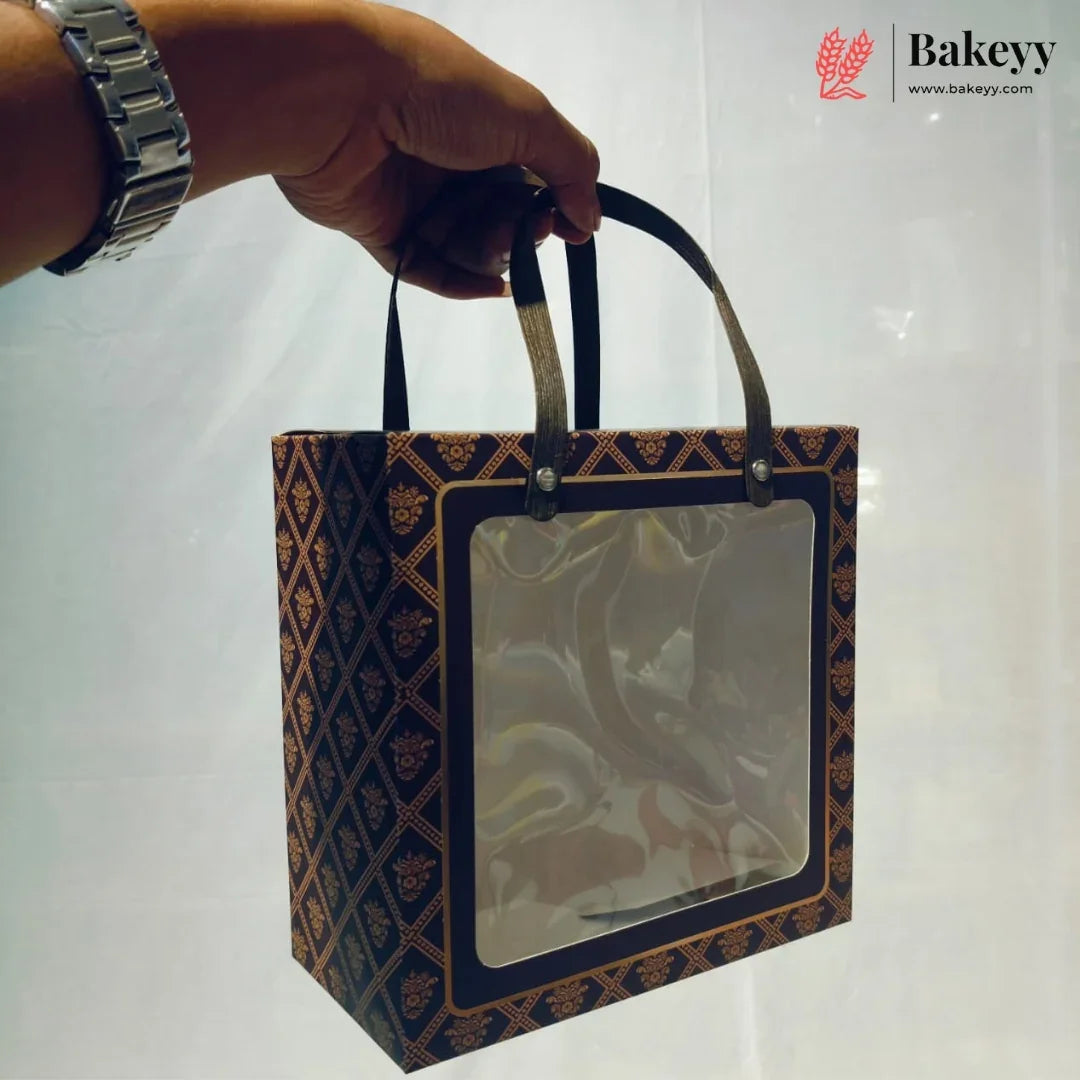 A hand holds a square bakery gift bag with a clear plastic window and dark handles. The bag has a dark navy blue and brown geometric patterned design.