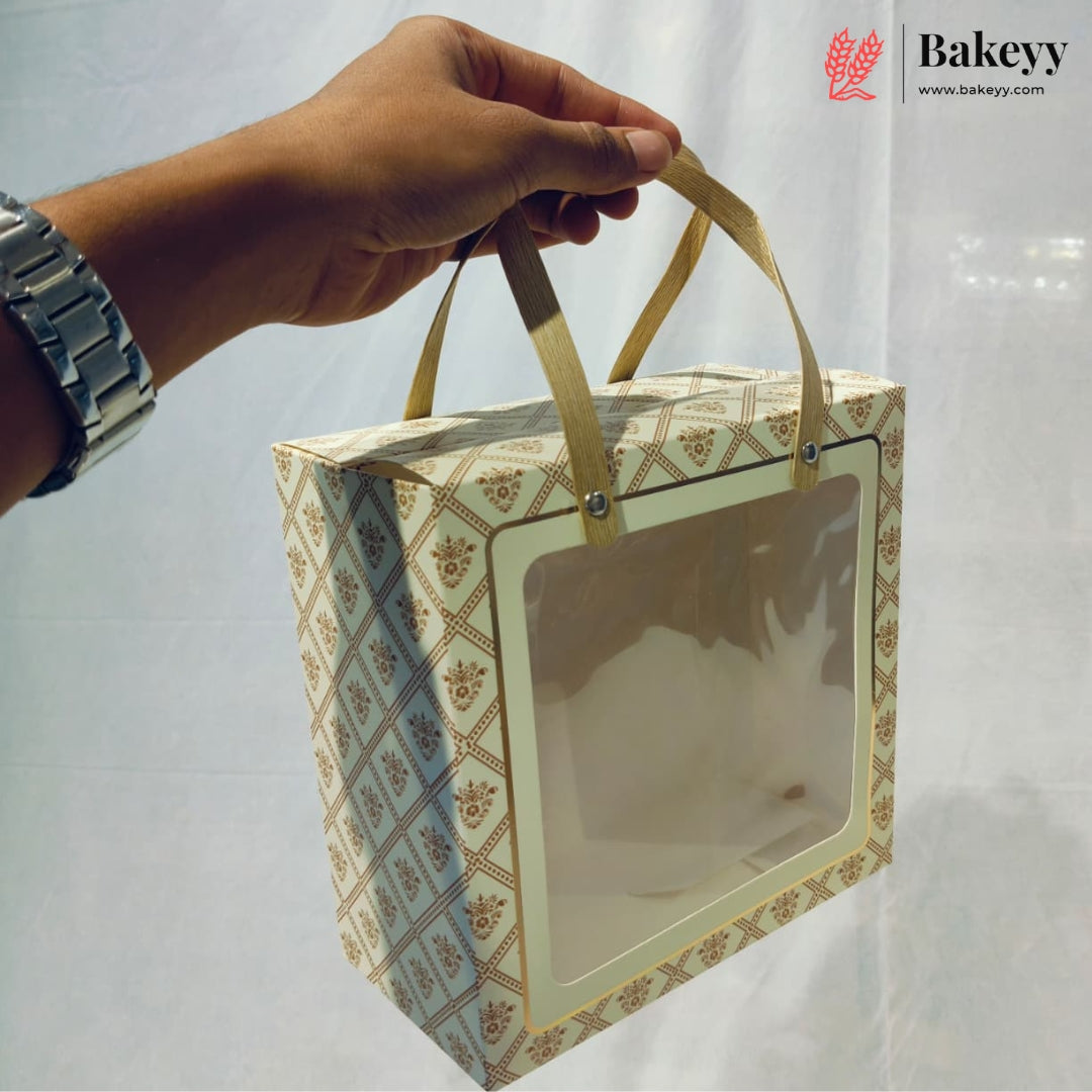 A hand holds a square bakery gift box with a clear front window and gold ribbon handles. The box has a beige and brown geometric pattern.