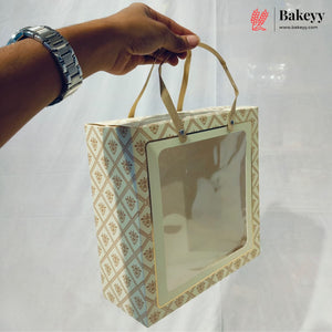 A hand wearing a silver watch holds a square bakery gift bag with a clear window and beige handles. The bag has a cream and gold geometric pattern, set against a white draped backdrop with a Bakeyy logo in the top right corner.