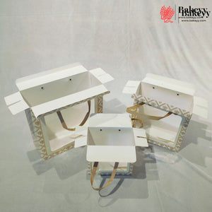 Three white bakery gift boxes with patterned sides and tan handles, displayed open on a light gray fabric backdrop. A bakery logo watermark is visible in the top right corner.