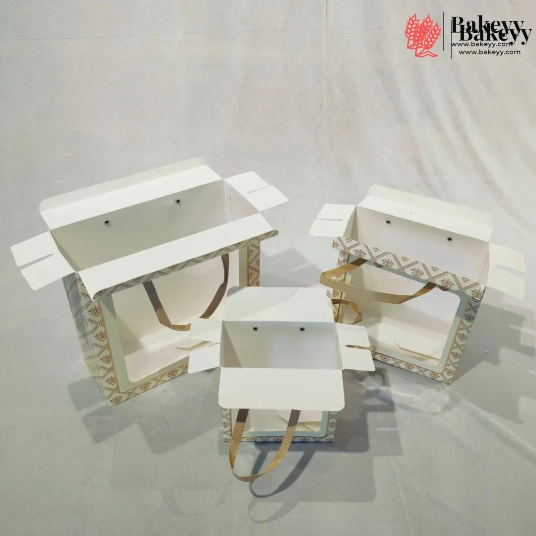 Three white bakery gift boxes with patterned sides and tan handles, displayed open on a light gray fabric backdrop. A bakery logo watermark is visible in the top right corner.
