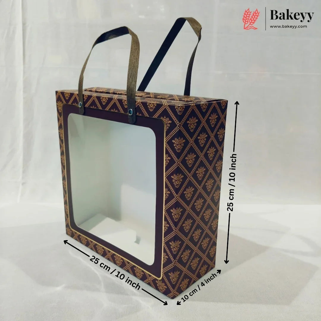 A square bakery gift box with a clear front window and two handles, shown with dimension arrows and labels indicating 25 cm by 25 cm by 10 cm. The box has a dark navy and gold geometric pattern.