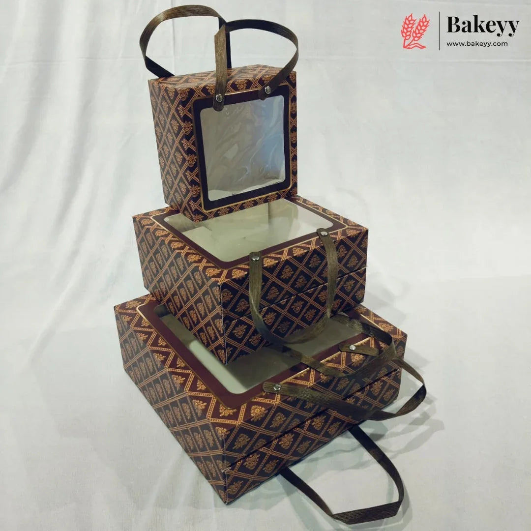 Three stacked rectangular bakery boxes with clear window lids and dark brown handles, featuring a repeating geometric pattern in brown and gold. The boxes are arranged on a white fabric backdrop, with a Bakeyy logo watermark in the top right corner.