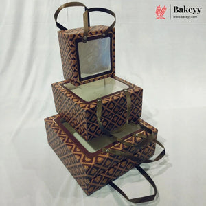 Three stacked rectangular bakery boxes with clear window lids and dark brown handles, featuring a repeating geometric pattern in brown and gold. The boxes are arranged on a white fabric backdrop, with a Bakeyy logo watermark in the top right corner.