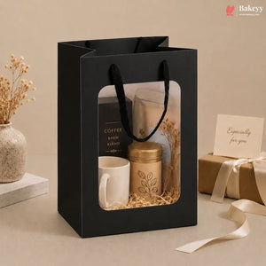 A black gift bag with a clear window displaying a coffee gift set, including a white mug, a gold-lidded tin, and a candle, set against a warm beige background with decorative props.