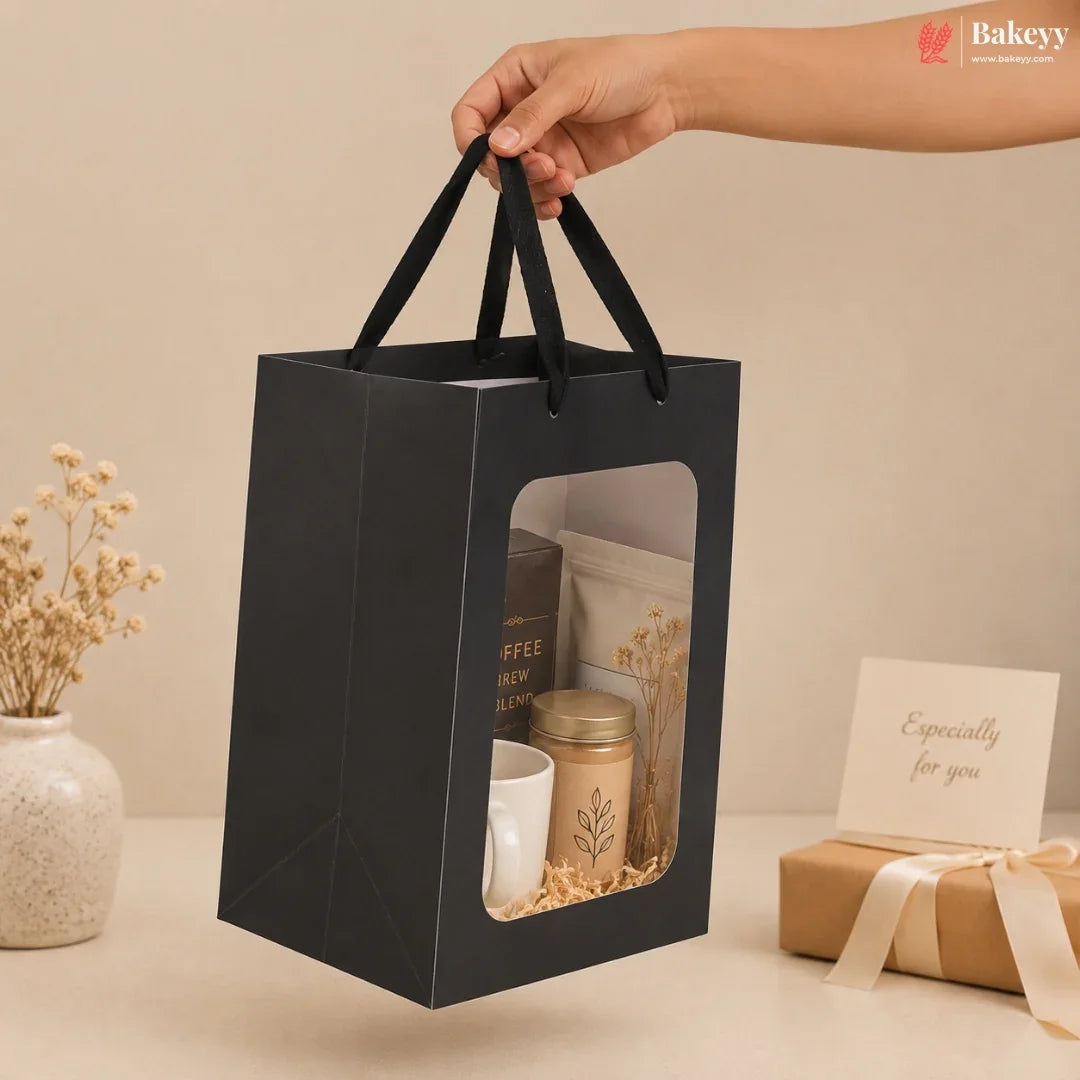 A black paper gift bag with a clear window and black handles, held by a hand. Inside the bag are coffee-related items including a jar, a mug, and a pouch.