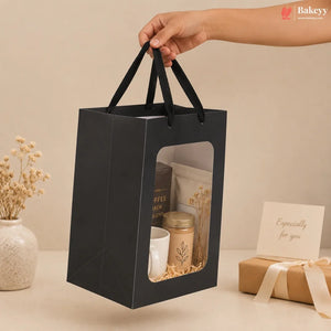 A black paper gift bag with a clear window and black handles, held by a hand. Inside the bag are coffee-related items including a jar, a mug, and a pouch.