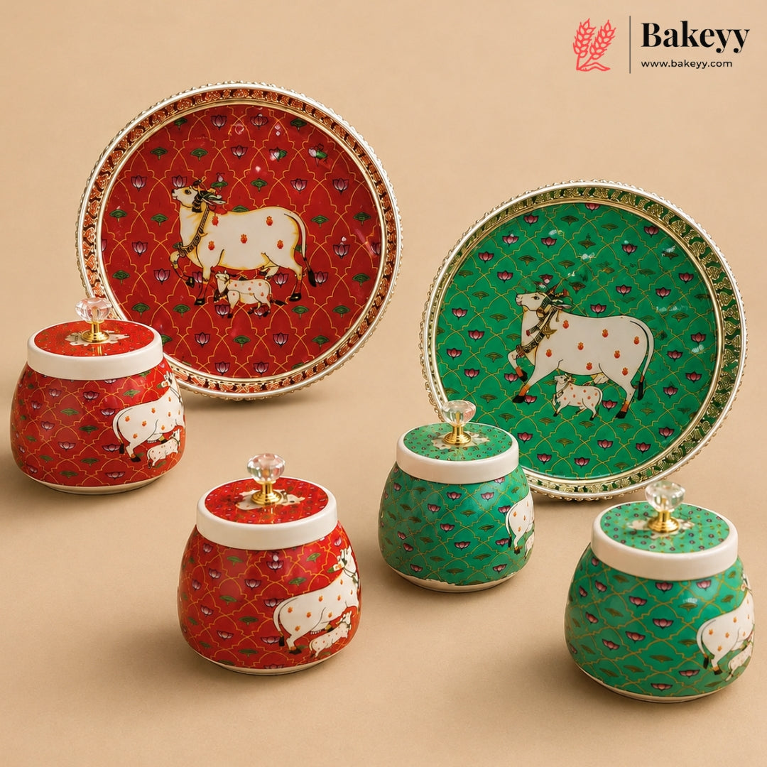 A set of decorative ceramic baking containers and serving plates from Bakeyy, featuring red and green enamel bodies with white cow illustrations and gold-toned lids with crystal knobs, displayed on a beige background.