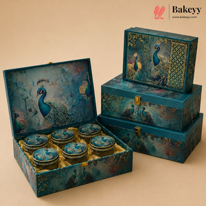 A decorative gift set of round tin jars and matching boxes featuring peacock illustrations in teal, blue, and gold tones, arranged on a warm beige background. One large box is open to show the six round tins inside, and two smaller matching boxes are stacked beside it.