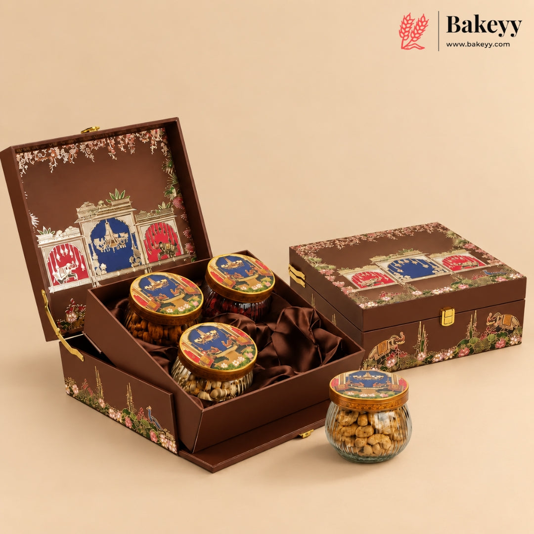 A premium chocolate gift set in an ornate brown suitcase-style box with floral and architectural illustrations, shown open with three round jars of chocolate and one glass jar in front. The brand Bakeyy is visible in the top right corner.