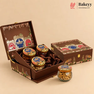 A premium chocolate gift set in an ornate brown suitcase-style box with floral and architectural illustrations, shown open with three round jars of chocolate and one glass jar in front. The brand Bakeyy is visible in the top right corner.