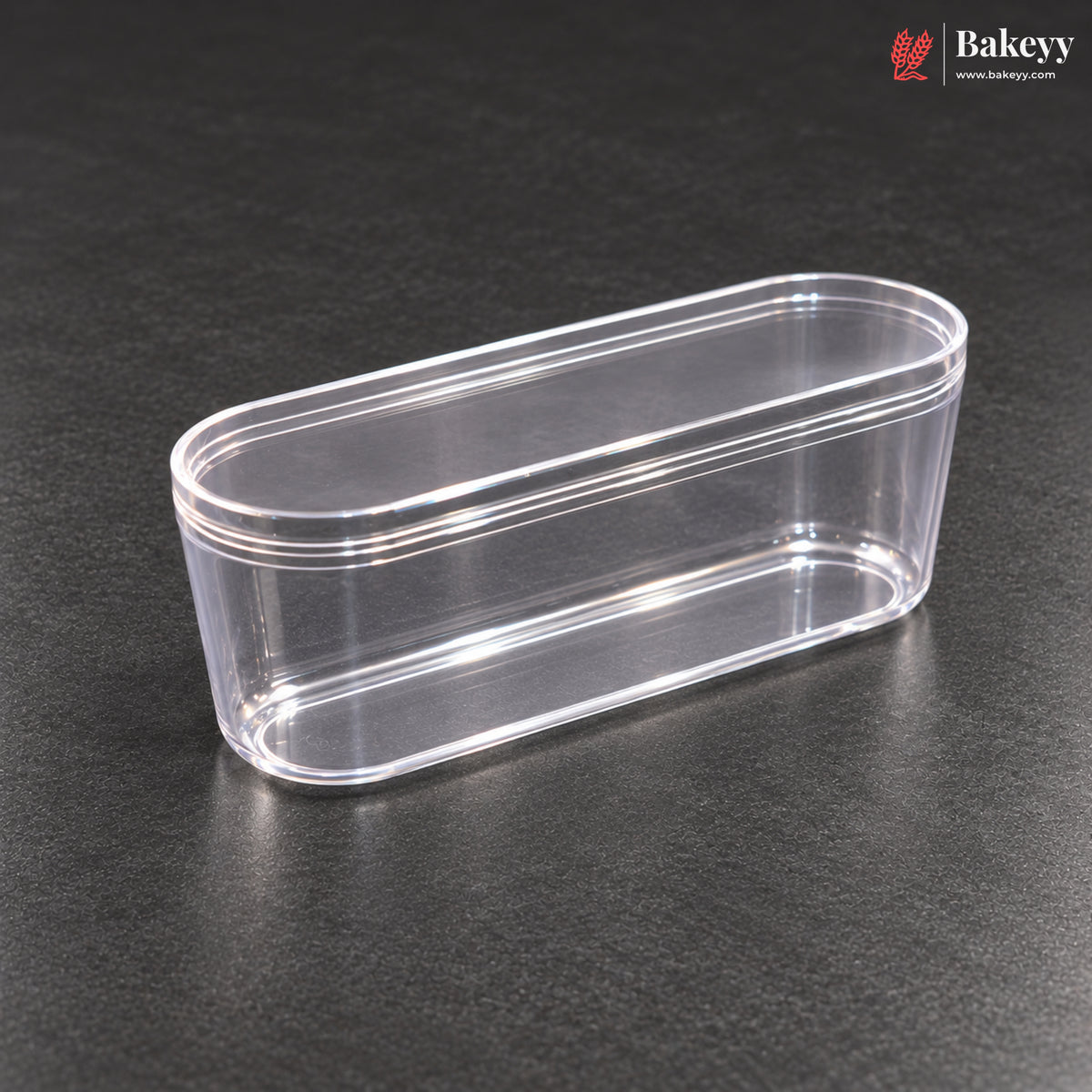 A clear plastic oval-shaped container with a matching lid, shown on a dark textured surface. The Bakeyy logo is visible in the top right corner.