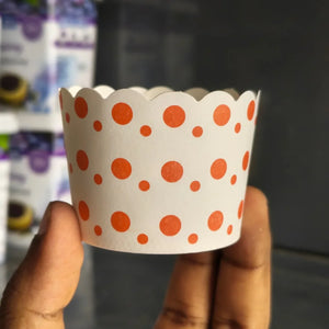A hand holding a white scalloped paper cupcake liner decorated with orange polka dots, with blurred product packaging in the background.