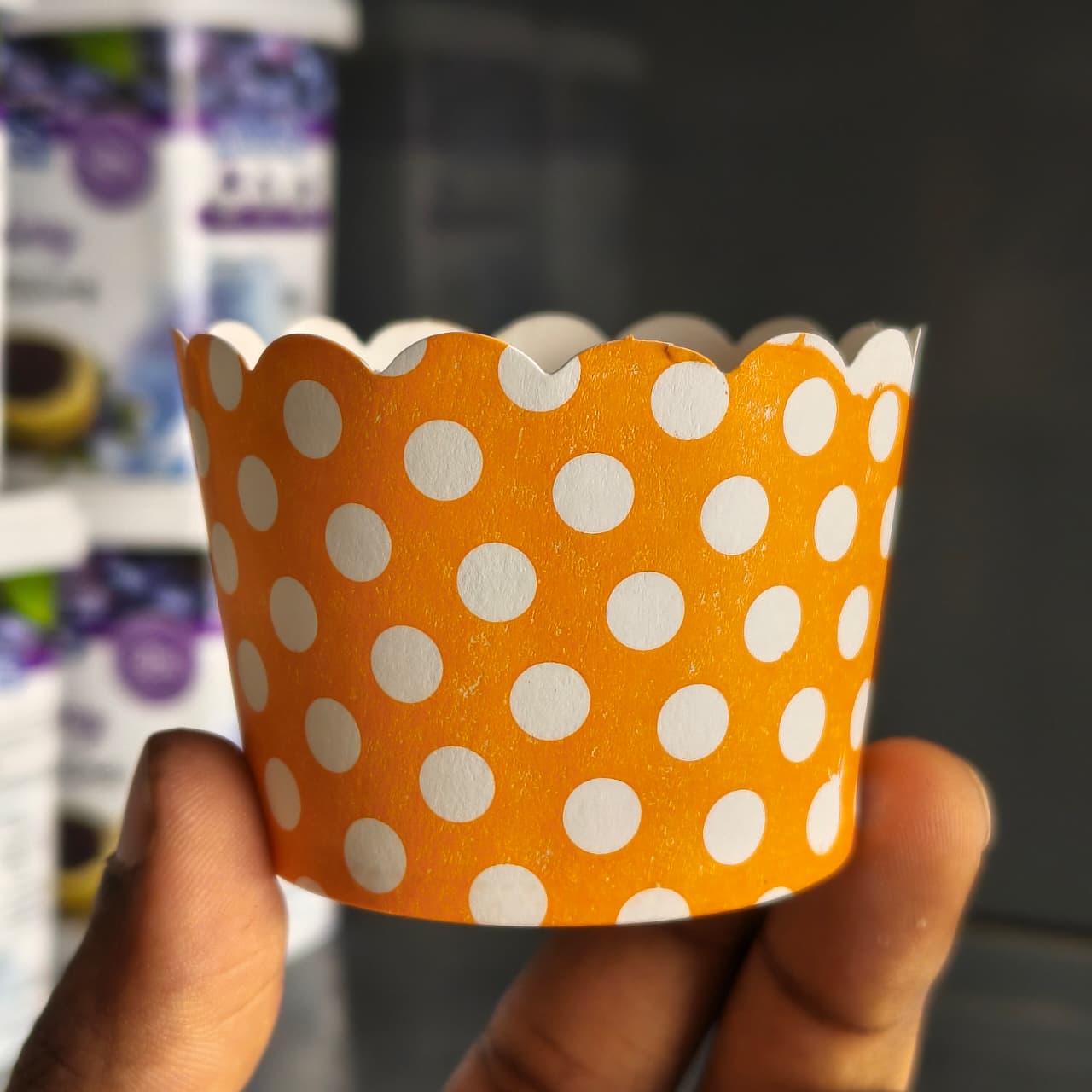An orange paper cupcake liner with white polka dots being held by a hand, with a blurred background showing shelves of packaged goods.