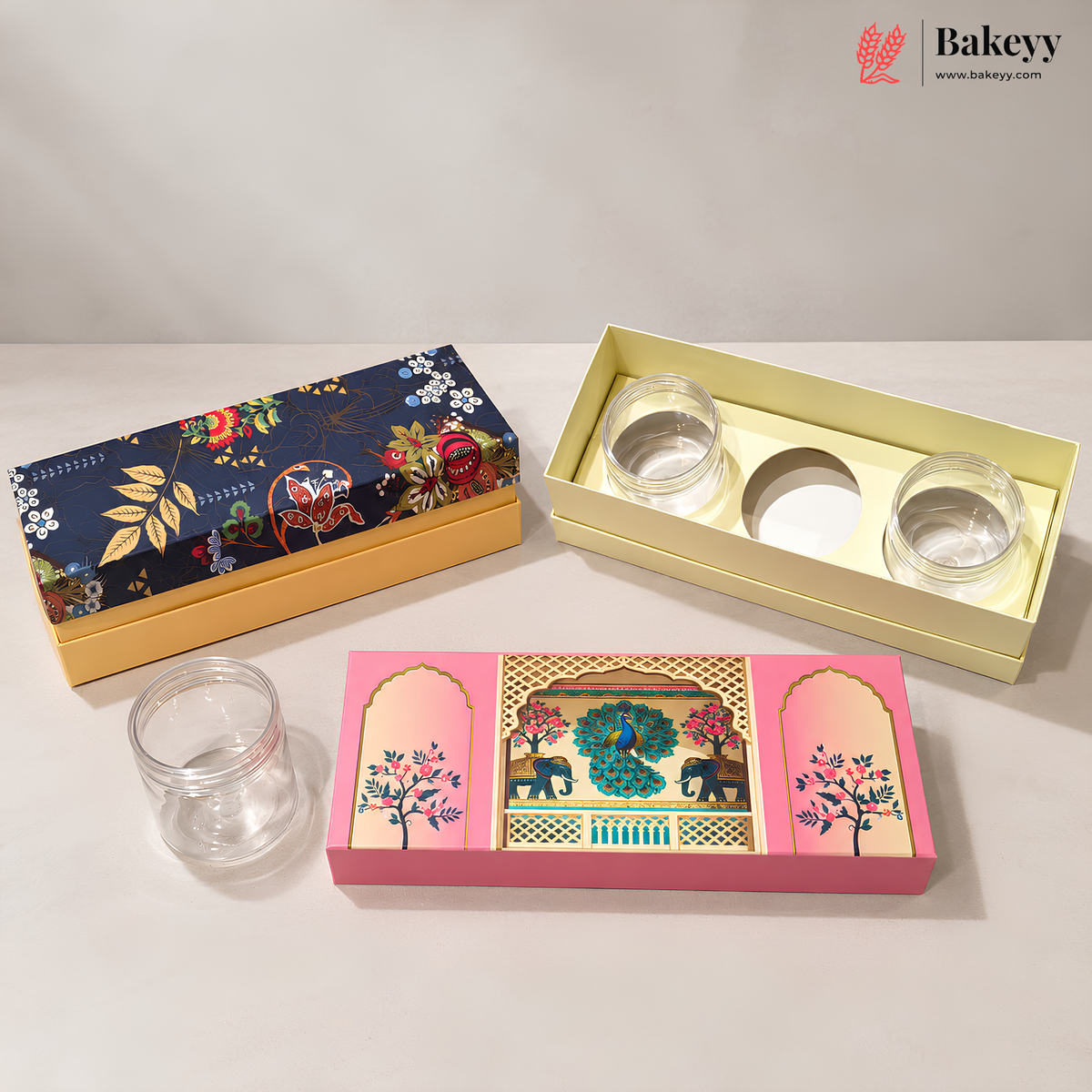 Three decorative glass jars in different packaging styles on a light gray surface: a navy floral box with gold base, an open pale yellow box with two clear glass jars, and a pink illustrated box with one empty clear glass jar.