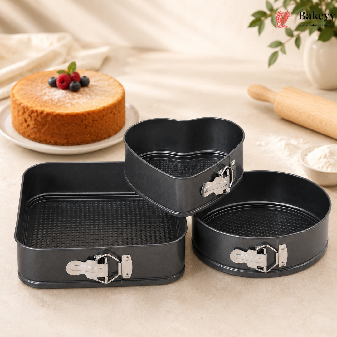 Three-piece black non-stick springform cake pan set with silver clamps, displayed on a beige fabric surface with baking props like a rolling pin, flour, and a decorated cake in the background.
