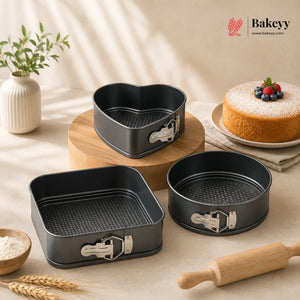 Three dark gray non-stick springform pans in heart, square, and round shapes are arranged on a beige tabletop with a wooden rolling pin, wheat stalks, a small vase with greenery, and a decorated cake in the background. The Bakeyy brand logo is visible in the top right corner.