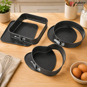 Black non-stick springform pan set on a wooden table, including a square pan with a matching base and two heart-shaped rings, with kitchen props like eggs, a whisk, and a plant in the background.