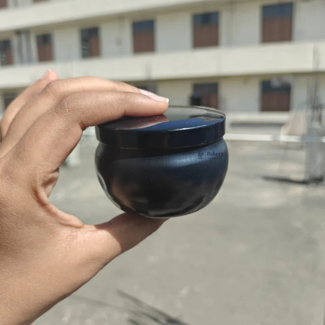 Hand holding a small black round jar container with Bakeyy logo, outdoor background