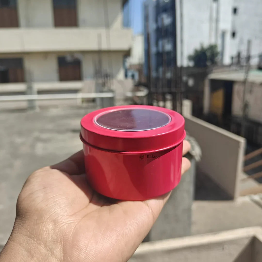 Hand holding a round pink metal tin container with Bakeyy branding, outdoors in sunlight.
