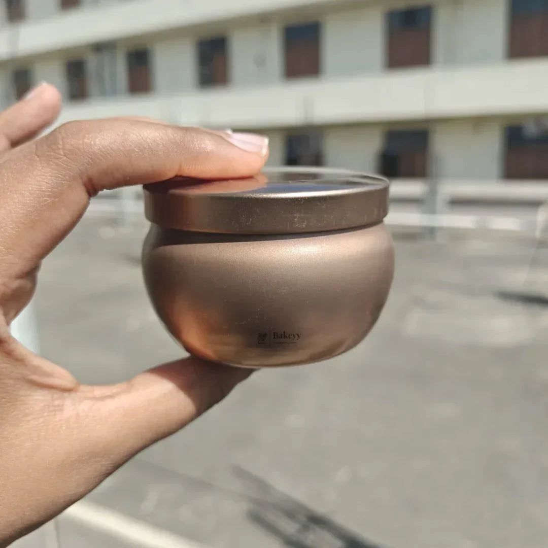 Hand holding a metallic round jar with Bakeyy logo, baking packaging product outdoors