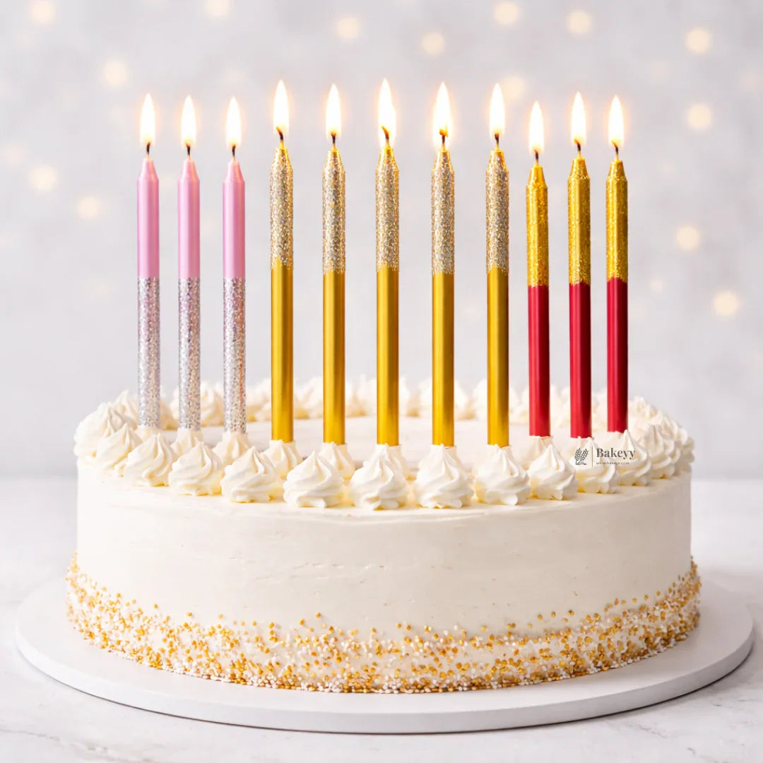 Decorative birthday cake with tall pink, gold, and red candles, white frosting, gold sprinkles, Bakeyy.