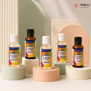 Four bottles of Bakeyville oil soluble flavours arranged on pastel display pedestals against a clean studio backdrop. The bottles include Makkurrim, Banana, Cheddar Cheese, and Peach flavours, with two amber glass bottles and two clear plastic bottles, each labeled with the Bakeyville logo.