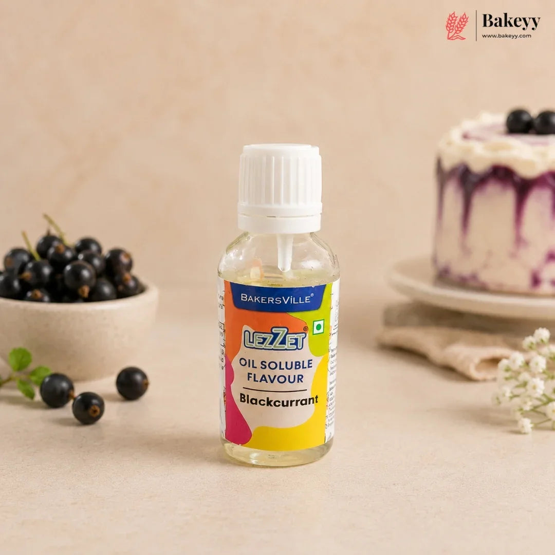 A bottle of Bakersville Uzley Blackcurrant Oil Soluble Flavour, set on a beige surface with fresh blackcurrants and a decorated cake in the background.