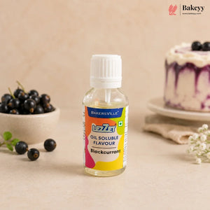 A bottle of Bakersville Uzley Blackcurrant Oil Soluble Flavour, set on a beige surface with fresh blackcurrants and a decorated cake in the background.