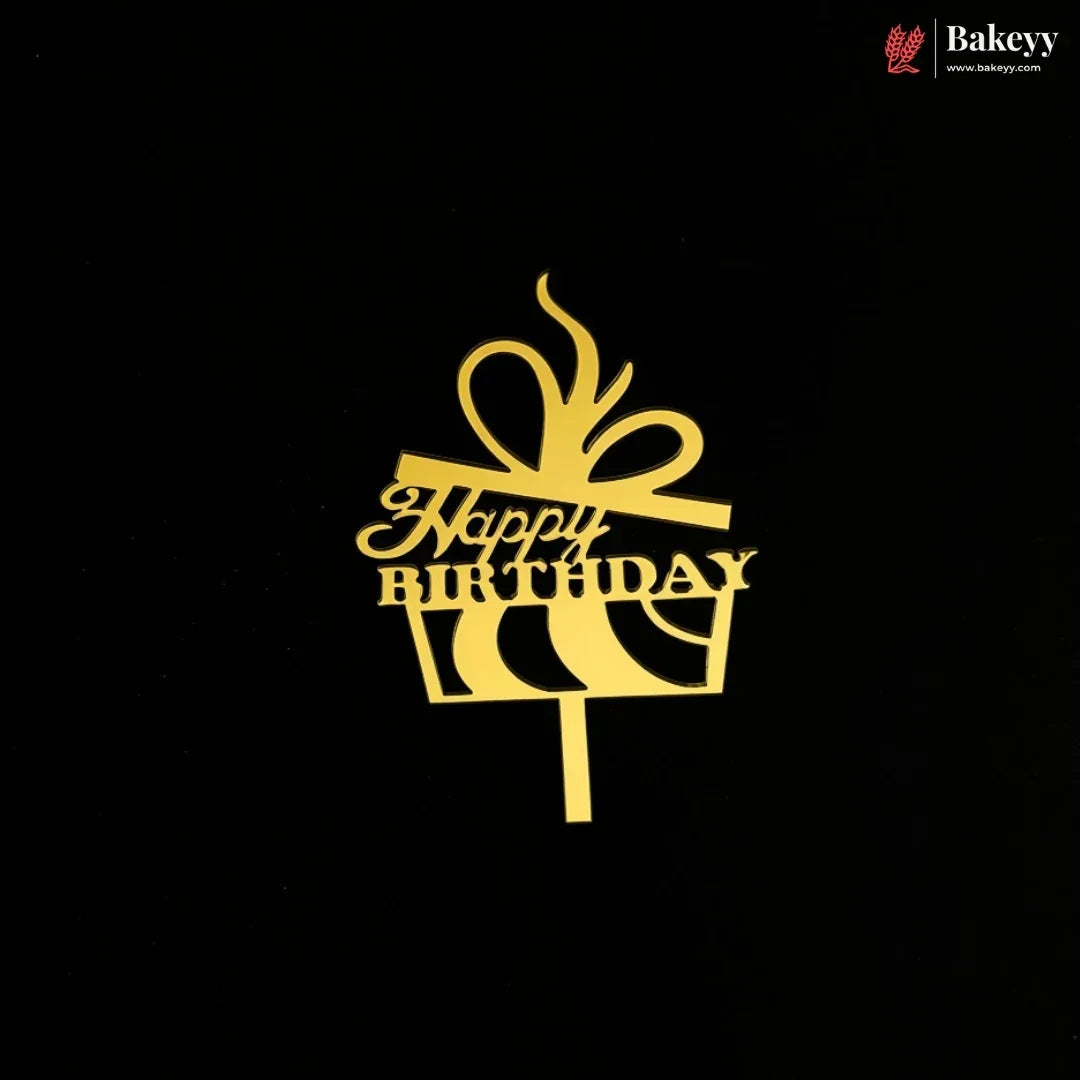 A gold cake topper shaped like a gift box with a bow and the words 'Happy Birthday' on a black background. A small Bakeey logo appears in the top right corner.