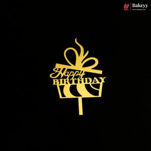 A gold cake topper shaped like a gift box with a bow and the words 'Happy Birthday' on a black background. A small Bakeey logo appears in the top right corner.