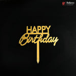 Gold metal 'HAPPY Birthday' cake topper with a vertical stick, displayed on a solid black background. A small 'Bakeyy' brand logo is visible in the top right corner.