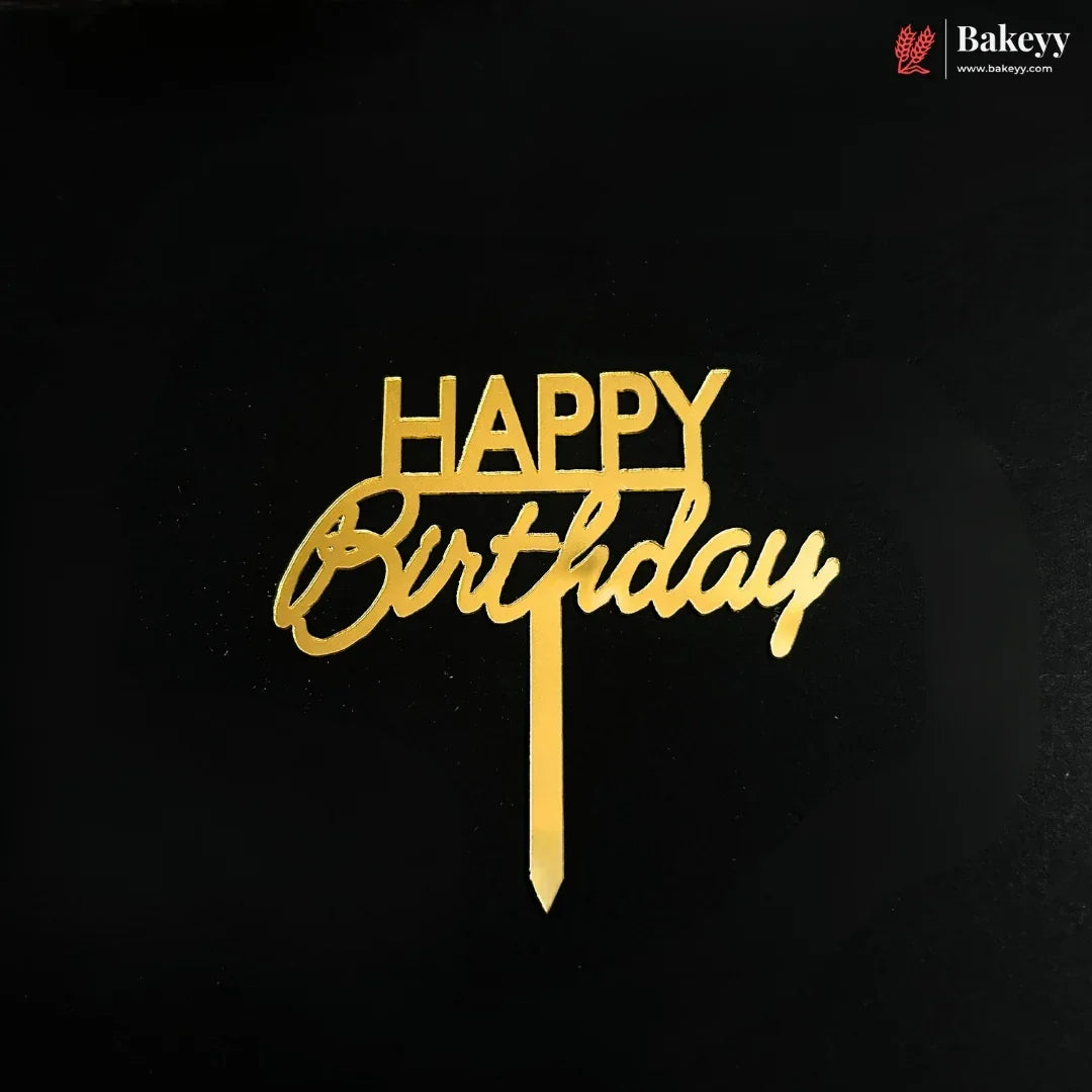 Gold metal 'HAPPY Birthday' cake topper with a vertical stick, displayed on a solid black background. A small 'Bakeyy' brand logo is visible in the top right corner.