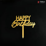 Gold metal 'HAPPY Birthday' cake topper with a vertical stick, displayed on a solid black background. A small 'Bakeyy' brand logo is visible in the top right corner.