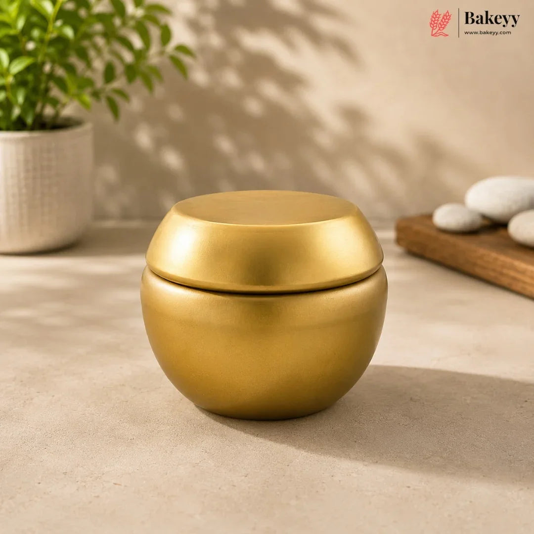 Gold-colored container on a beige surface with a plant and decorative items in the background, featuring the Bakeyy brand.