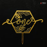 A gold metallic hexagonal cake topper with honeycomb patterns, bees, and the word 'one' in script, shown on a black background. A Bakeey watermark is visible in the top right corner.