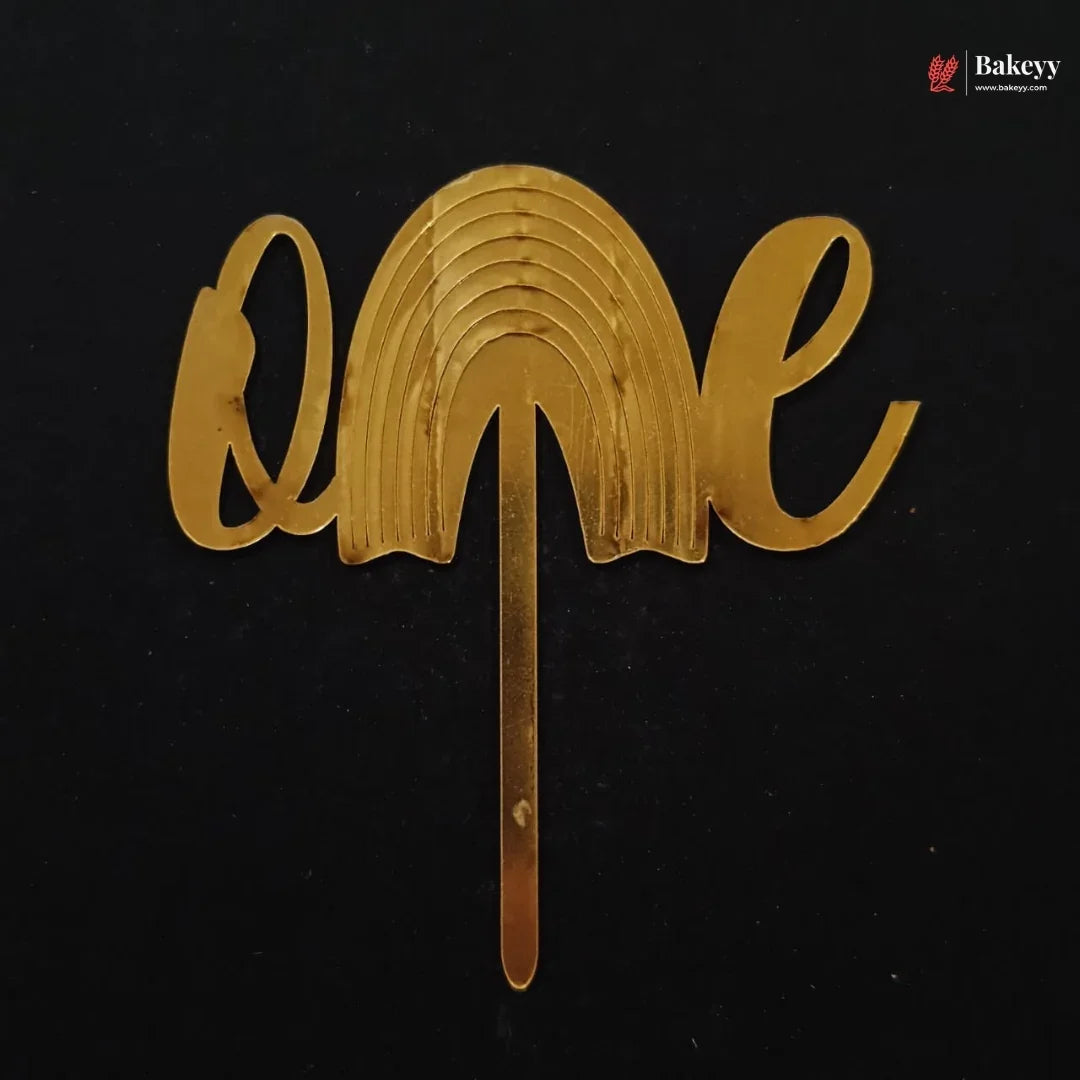 A gold metallic 'one' cake topper with a rainbow arch design and a stick base, shown on a black background with a Bakeey watermark in the top right corner.