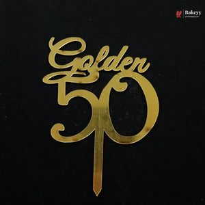 A gold metallic 'Golden 50' cake topper with a single wire stake, shown on a solid black background. A small 'Bakeyy' brand watermark is visible in the top right corner.