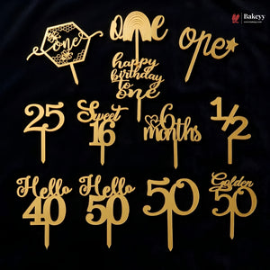 A set of gold laser-cut cake toppers displayed on a black background, including designs for one, twenty-five, thirty, thirty-one, forty, fifty, sixty, seventy, eighty, ninety, and hundred years, as well as 'one happy birthday one' and 'one open'. A Bakeyy logo is visible in the top right corner.