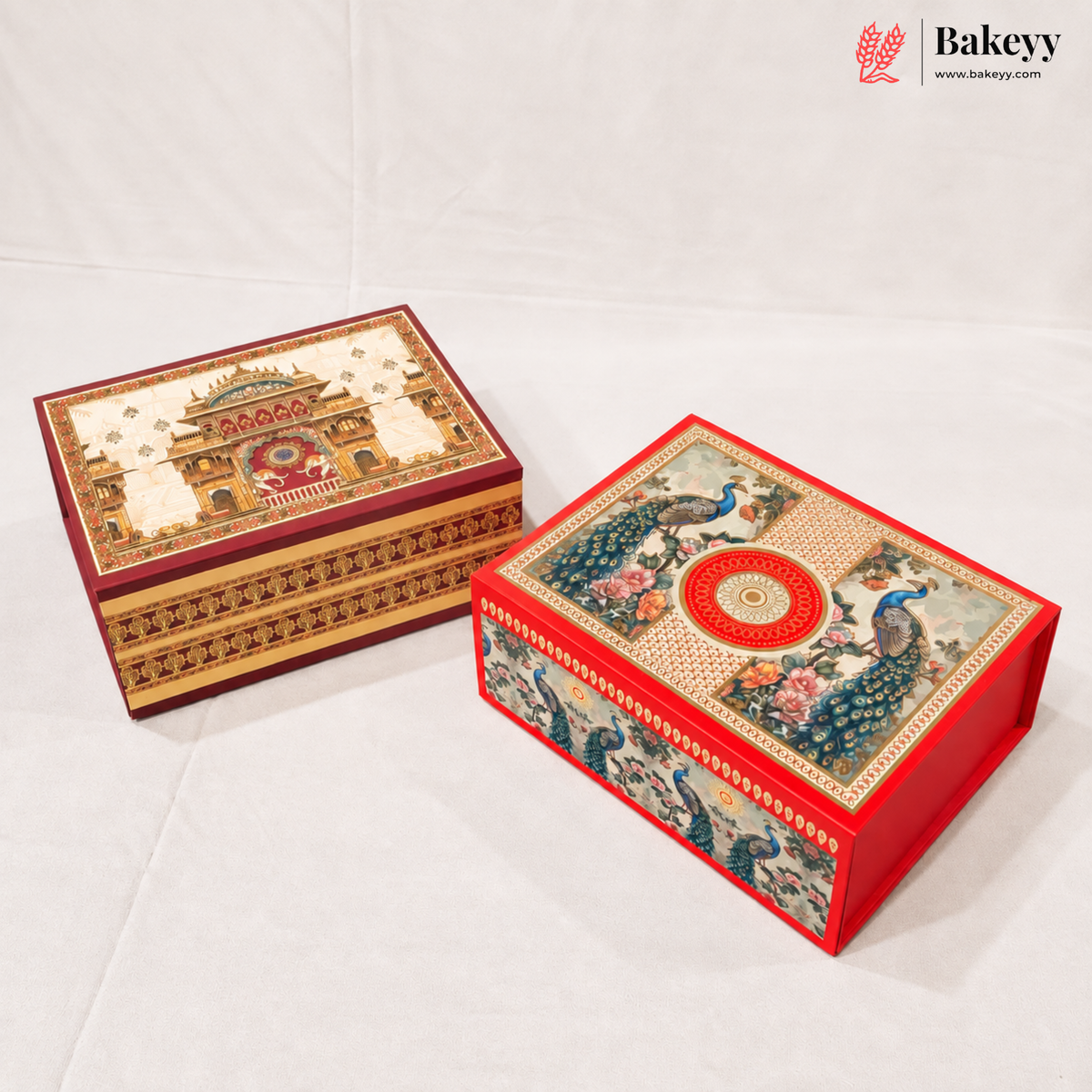 Two decorative rectangular gift boxes with ornate printed designs, one featuring a temple illustration and the other peacocks, displayed on a white fabric background. A Bakeyy logo is visible in the top right corner.