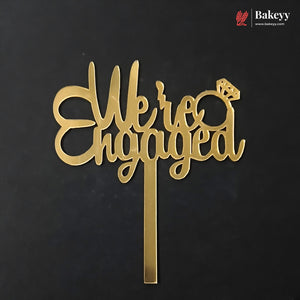 Gold metal 'We are Engaged' cake topper with a ring icon, shown on a black background with a Bakeey brand watermark in the top right corner.